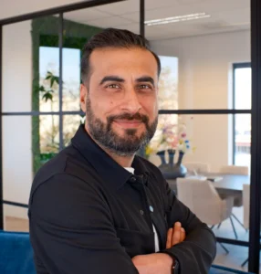 Man met verzorgde baard en zwart overhemd staat met armen gekruist voor glazen wanden en kantoorinrichting