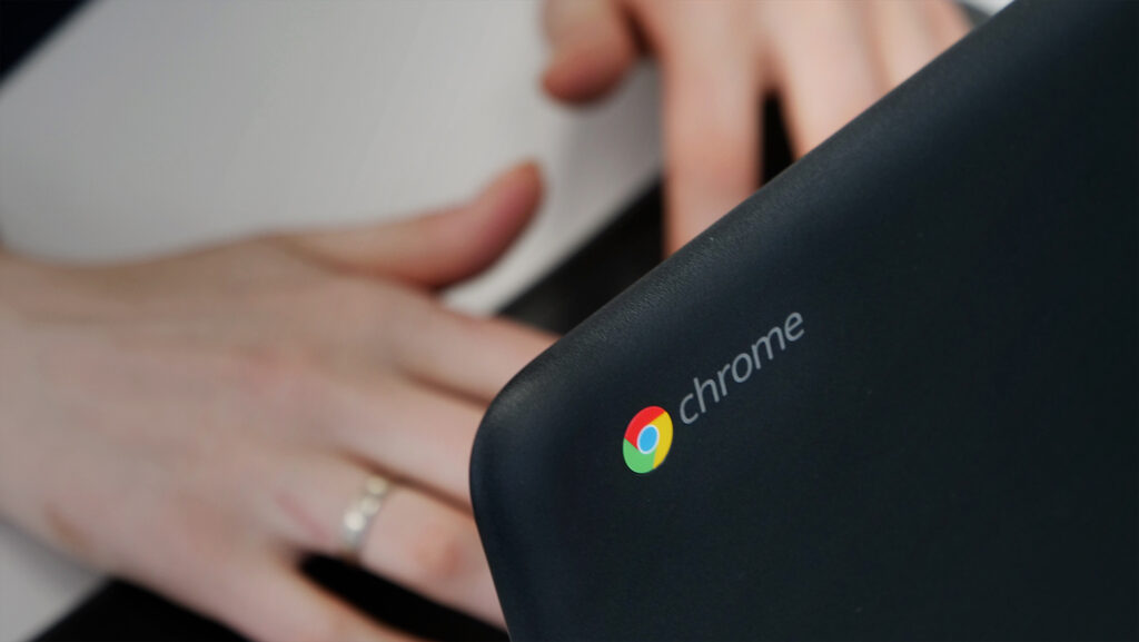 Een deel van een zwarte laptop met kleurrijk Chrome-logo en handen op een tafel eronder