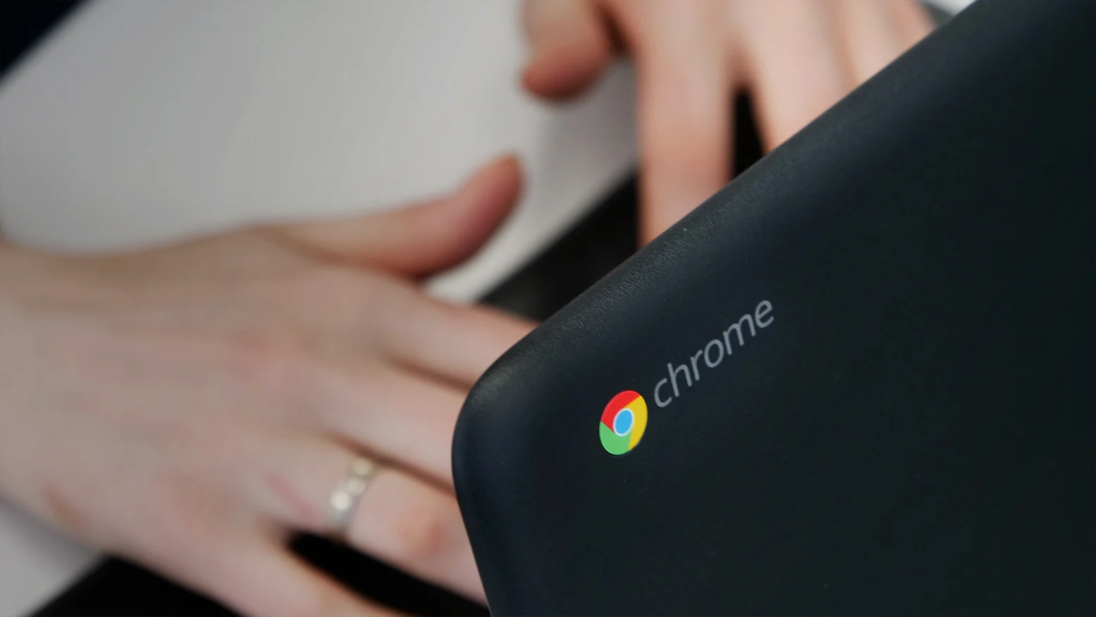 Een deel van een zwarte laptop met kleurrijk Chrome-logo en handen op een tafel eronder
