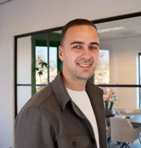 Man in een donkere jas en wit T-shirt staat glimlachend in een lichte kantoorruimte met grote ramen