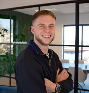 Een jonge man met lichtbruin haar en baard staat met armen gekruist in een modern kantoor.