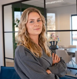 Vrouw met lang golvend blond haar en grijze outfit staat met armen over elkaar in modern kantoor.