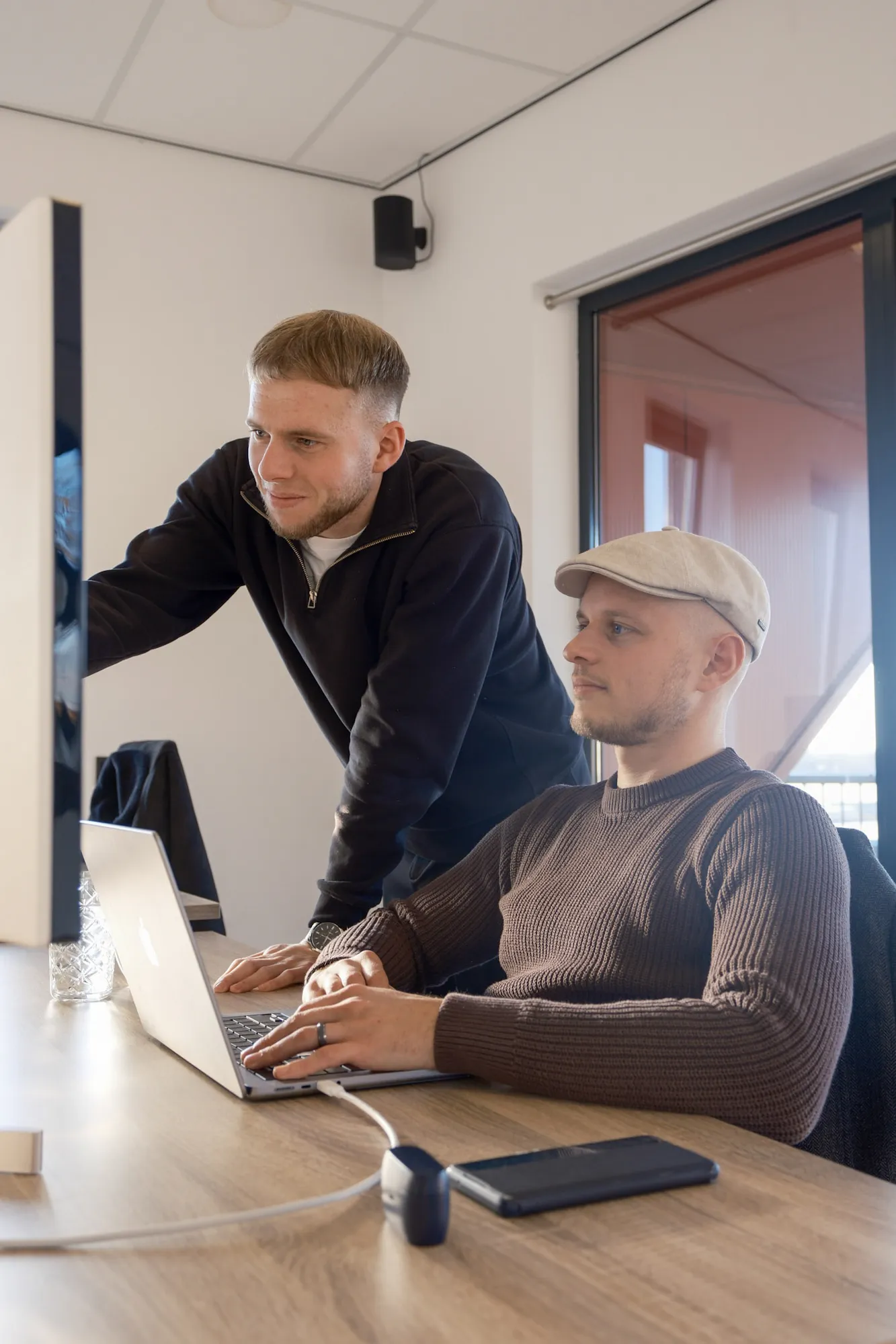 Twee mannen werken samen aan een bureau met laptop en computer in een lichte kantoorruimte.