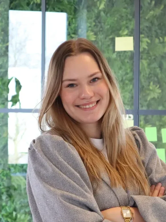 Een jonge vrouw met lange golvende haren staat in een lichte ruimte met grijze trui en spijkerbroek.
